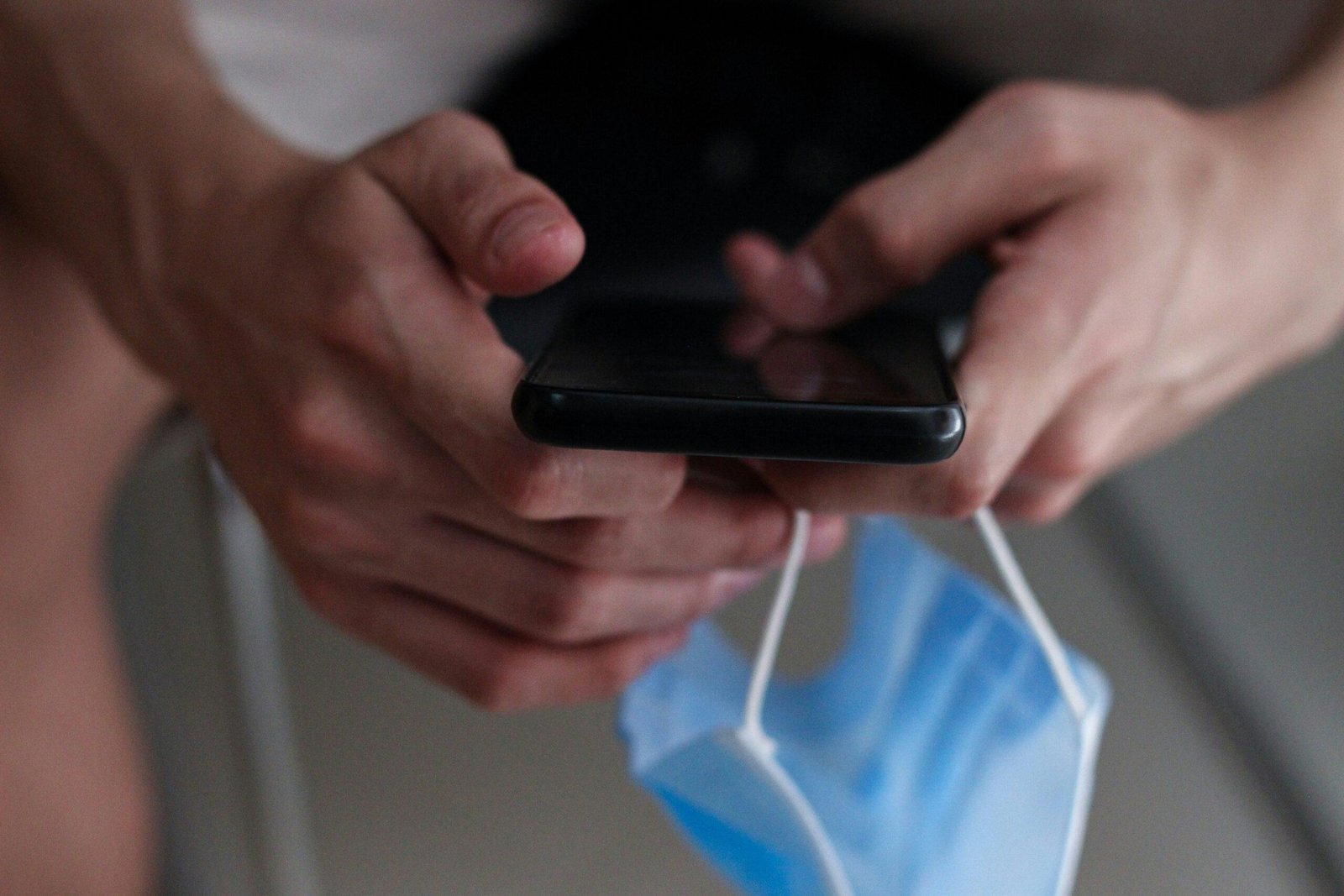 Manos sosteniendo un celular y una mascarilla quirúrgica azul