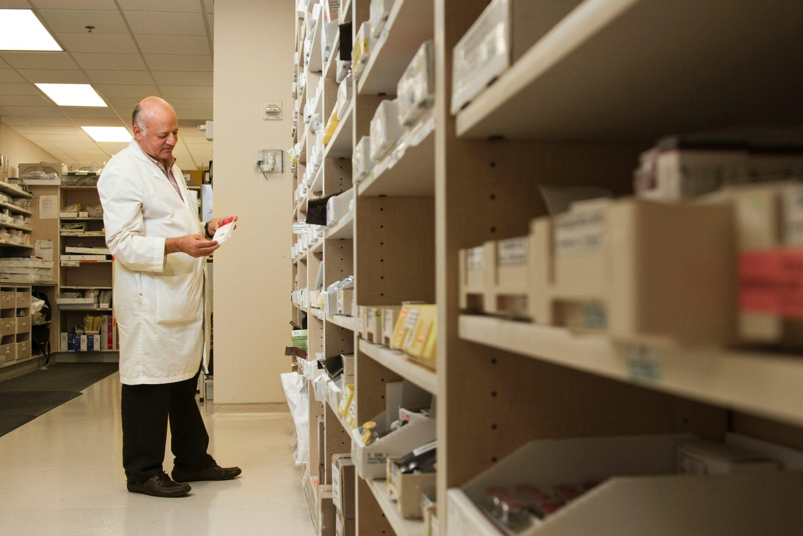 Farmacéutico con bata blanca revisando la caja de un medicamento junto a los estantes de una farmacia
