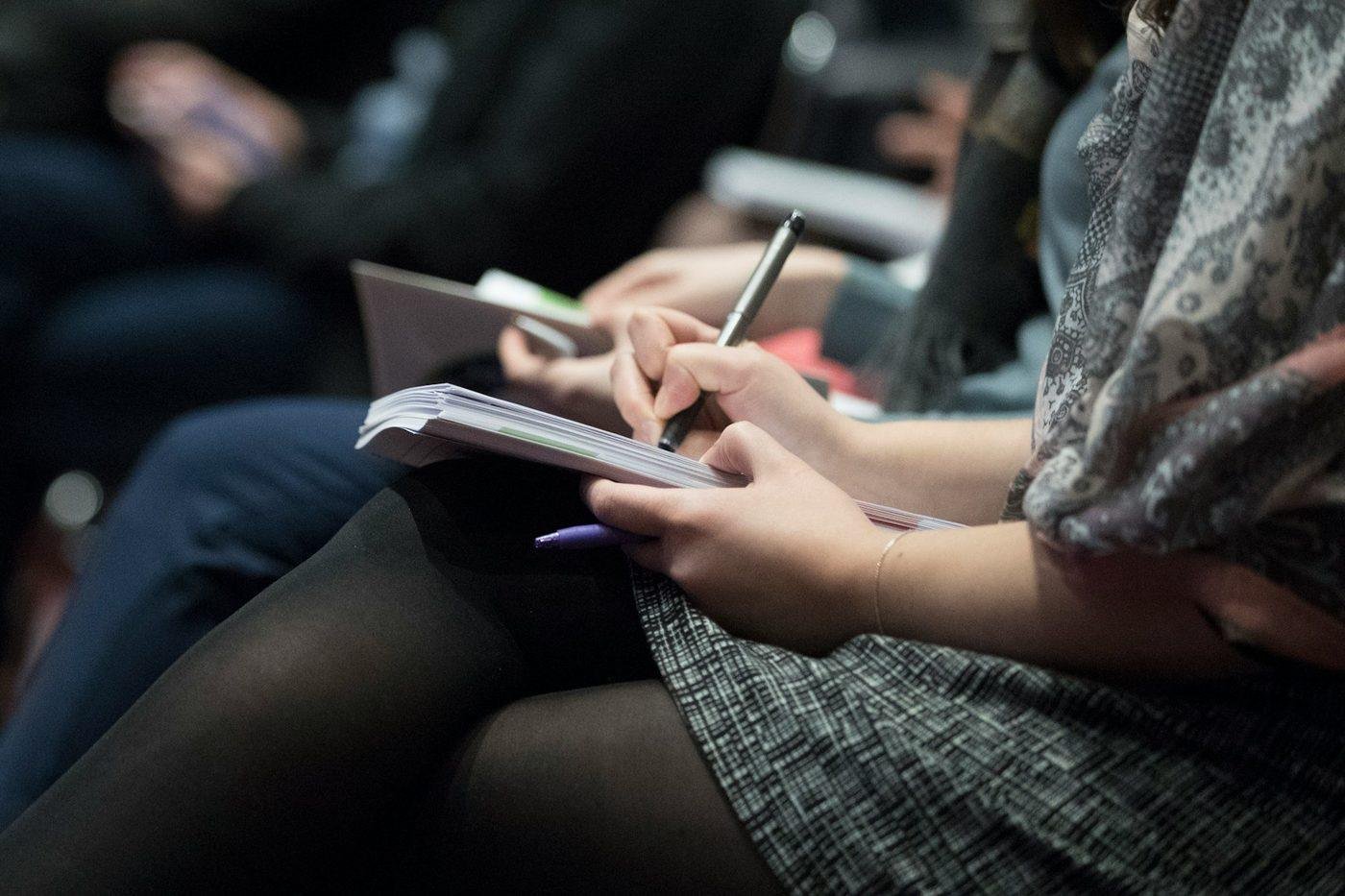 Mano escribiendo en una libreta durante una conferencia, con otros asistentes tomando notas al fondo